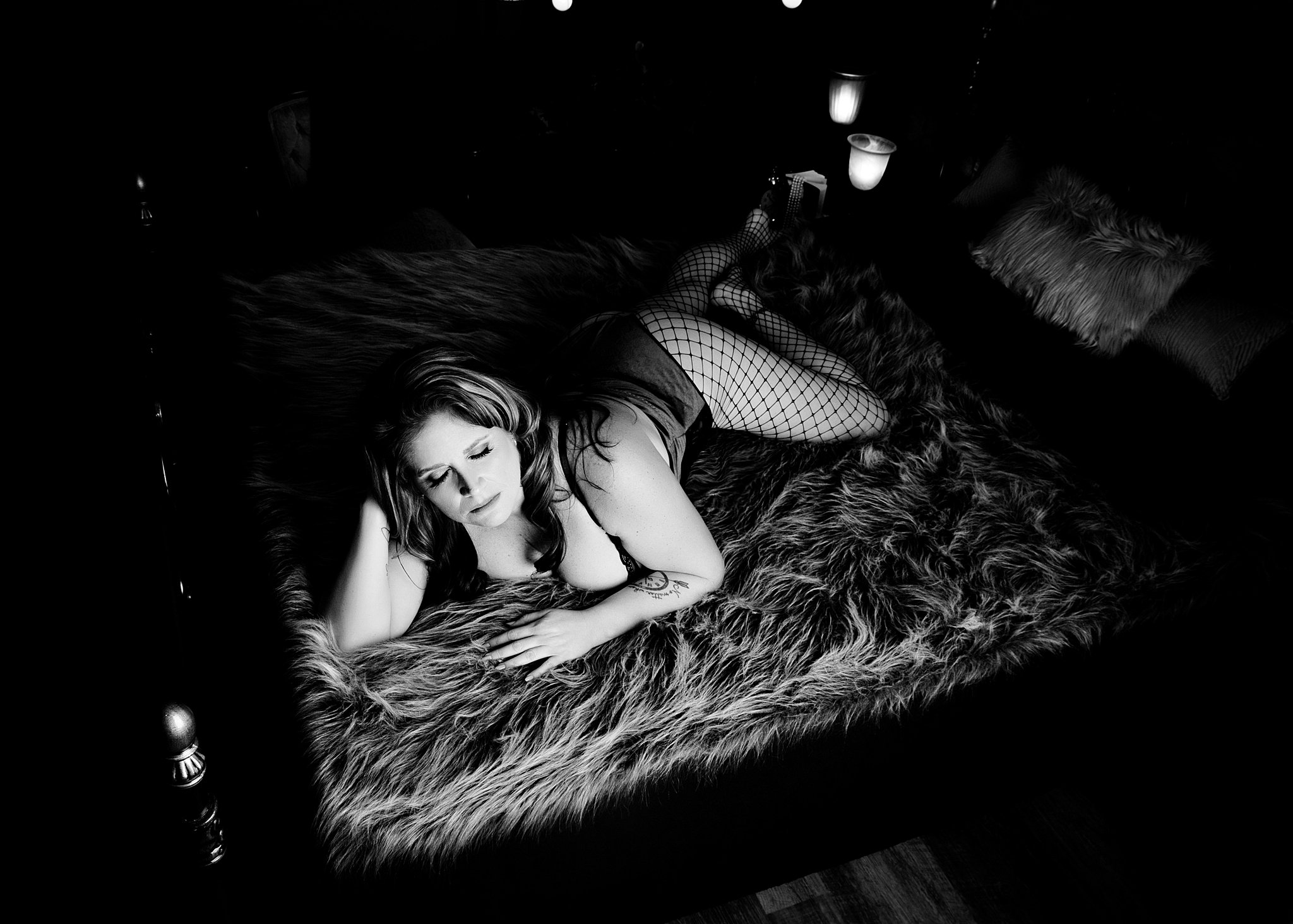 A woman in fishnets and lingerie lays across a bed in a studio on a fur blanket